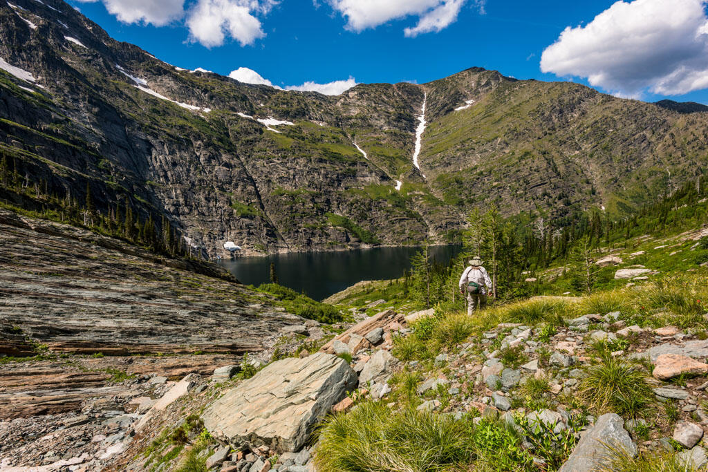an image of a lake, surrounded by steep rocks/mountains. the sky is blue and mostly clear and there are several kinds of green trees in the foreground.