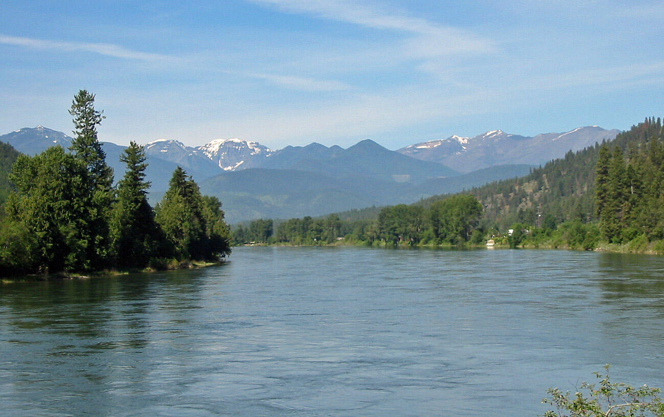 a picturesque image of a shallow creek, with grassy banks and trees and mountains in the background